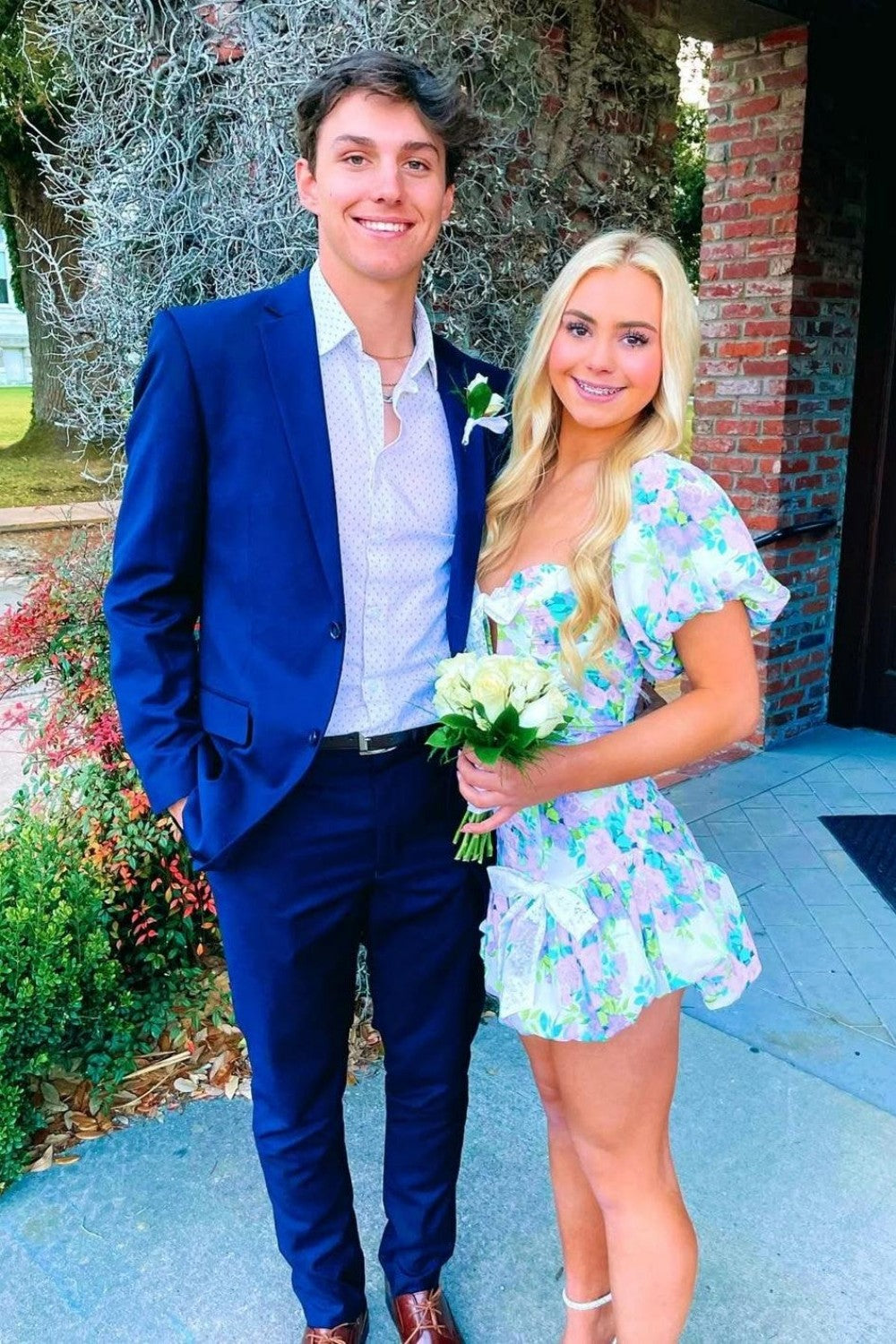 Man in a blue suit and woman in a floral dress standing outdoors.